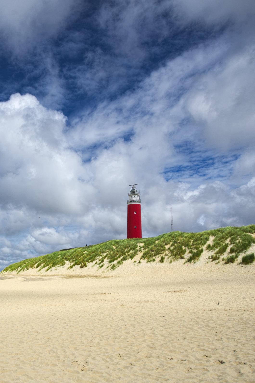 Een strand met de roden vuurtoren van Texel in de duinen en een blauwe lucht met grote witte wolken.