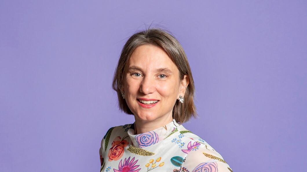 Foto van Laura Bromet, een witte vrouw. Ze draagt een wit shirt met bloemetjes in verschillende kleuren, heeft donkerblond haar en lacht.