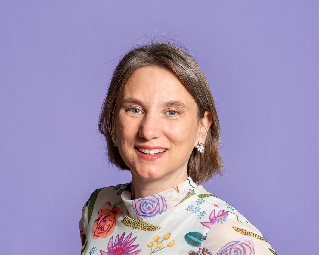 Foto van Laura Bromet, een witte vrouw. Ze draagt een wit shirt met bloemetjes in verschillende kleuren, heeft donkerblond haar en lacht.